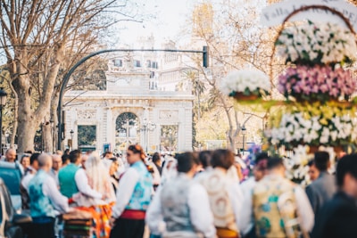 A group of people wearing traditional costumes gathers in front of an ornate stone archway. The scene is bustling with bright floral decorations and trees with leaves turning brown, suggesting an outdoor celebration or festival.
