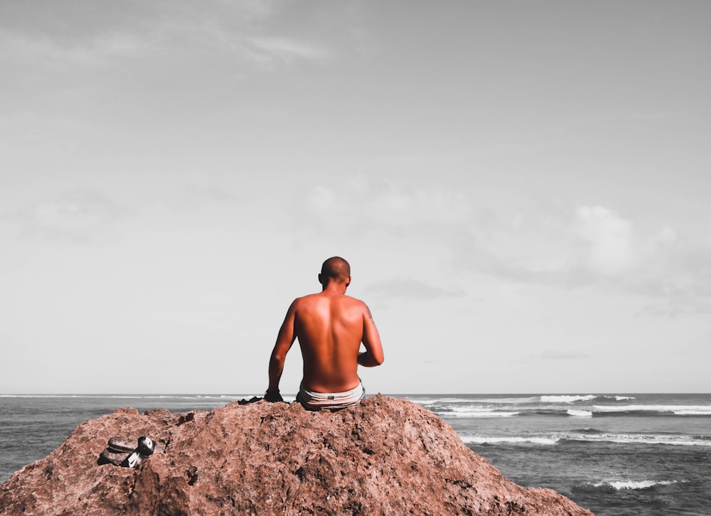 topless man sitting on rock near shore