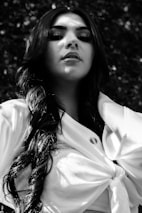 A person with long, wavy hair is looking upwards with a confident expression. They are wearing a light-colored top with a knot in the front, and the image is in black and white. The background is out of focus, consisting of foliage and natural elements.