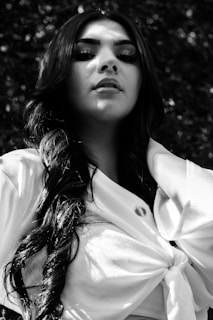 A person with long, wavy hair is looking upwards with a confident expression. They are wearing a light-colored top with a knot in the front, and the image is in black and white. The background is out of focus, consisting of foliage and natural elements.