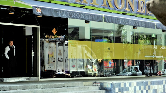A person wearing formal attire stands at the entrance of a building with large glass windows. The facade is modern with a prominent sign displaying 'Patrones'. The interior is partially visible and appears to be a commercial establishment. The exterior includes decorative elements and reflective surfaces, creating an upscale ambiance.