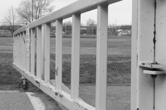 A black and white image depicting a close-up view of a metal gate or barrier on wheels, positioned on a paved surface. In the background, there are leafless trees and a field with some structures and a fence visible in the distance.