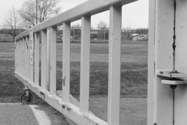 A black and white image depicting a close-up view of a metal gate or barrier on wheels, positioned on a paved surface. In the background, there are leafless trees and a field with some structures and a fence visible in the distance.