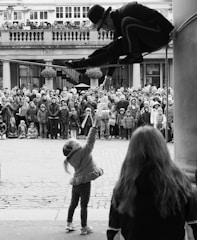 A man performs a high-wire act, balancing on a rope between two columns. He is dressed in a suit and hat and is interacting with a young girl reaching up to him. A large crowd gathers in the background, watching the performance with interest.