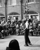 A performer balances a bicycle vertically on his chin in front of an audience seated and standing in an open courtyard. The onlookers, including adults and children, appear engaged and impressed by the stunt.