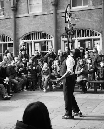 A performer balances a bicycle vertically on his chin in front of an audience seated and standing in an open courtyard. The onlookers, including adults and children, appear engaged and impressed by the stunt.