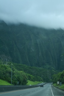 A highway curves gently through a lush, green landscape with dense vegetation and tall trees. A mountain with steep ridges is partially shrouded in thick, low-hanging clouds, creating a dramatic natural backdrop. Several cars are scattered along the road, suggesting a peaceful drive through a scenic area.