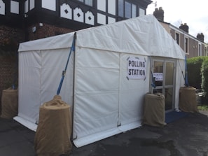 A white tent is set up outdoors, serving as a temporary polling station. Signs reading 'Polling Station' are displayed on the structure. The tent is secured with ropes and weights wrapped in brown coverings. The surrounding area includes a paved surface and nearby residential buildings.