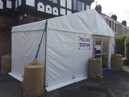 A white tent is set up outdoors, serving as a temporary polling station. Signs reading 'Polling Station' are displayed on the structure. The tent is secured with ropes and weights wrapped in brown coverings. The surrounding area includes a paved surface and nearby residential buildings.