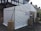 A white tent is set up outdoors, serving as a temporary polling station. Signs reading 'Polling Station' are displayed on the structure. The tent is secured with ropes and weights wrapped in brown coverings. The surrounding area includes a paved surface and nearby residential buildings.