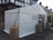A white tent is set up outdoors, serving as a temporary polling station. Signs reading 'Polling Station' are displayed on the structure. The tent is secured with ropes and weights wrapped in brown coverings. The surrounding area includes a paved surface and nearby residential buildings.