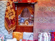 Intricate and colorful woven rugs and textiles are displayed, featuring geometric patterns in a variety of colors. They are arranged against an ornately carved wooden window or partition, behind which various decorative items, including vases and lamps, can be seen.