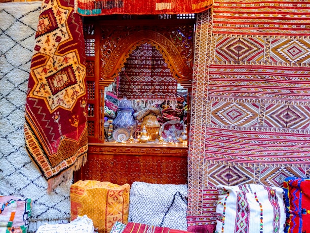 Intricate and colorful woven rugs and textiles are displayed, featuring geometric patterns in a variety of colors. They are arranged against an ornately carved wooden window or partition, behind which various decorative items, including vases and lamps, can be seen.