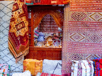 Intricate and colorful woven rugs and textiles are displayed, featuring geometric patterns in a variety of colors. They are arranged against an ornately carved wooden window or partition, behind which various decorative items, including vases and lamps, can be seen.