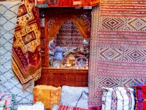Intricate and colorful woven rugs and textiles are displayed, featuring geometric patterns in a variety of colors. They are arranged against an ornately carved wooden window or partition, behind which various decorative items, including vases and lamps, can be seen.
