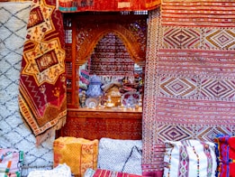 Intricate and colorful woven rugs and textiles are displayed, featuring geometric patterns in a variety of colors. They are arranged against an ornately carved wooden window or partition, behind which various decorative items, including vases and lamps, can be seen.