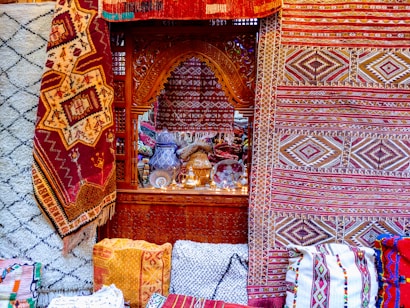Intricate and colorful woven rugs and textiles are displayed, featuring geometric patterns in a variety of colors. They are arranged against an ornately carved wooden window or partition, behind which various decorative items, including vases and lamps, can be seen.
