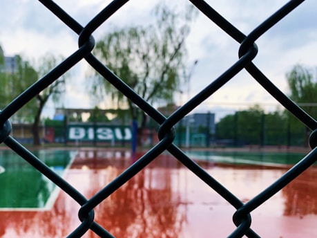 A chain-link fence in sharp focus with the background blurred, displaying a sports court with green and red surfaces. Trees and a building are visible in the distance, and the ground appears wet from recent rain.