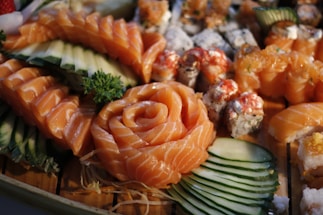 A beautifully arranged selection of sushi featuring fresh salmon slices in a rose pattern, accompanied by various rolls and garnished with cucumber slices and a sprig of parsley. The sushi pieces exhibit vibrant colors and intricate presentation on a wooden surface.