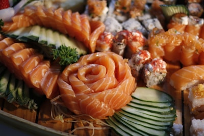 A beautifully arranged selection of sushi featuring fresh salmon slices in a rose pattern, accompanied by various rolls and garnished with cucumber slices and a sprig of parsley. The sushi pieces exhibit vibrant colors and intricate presentation on a wooden surface.
