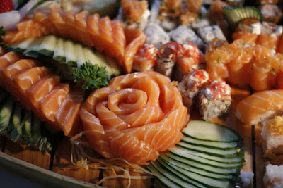 A beautifully arranged selection of sushi featuring fresh salmon slices in a rose pattern, accompanied by various rolls and garnished with cucumber slices and a sprig of parsley. The sushi pieces exhibit vibrant colors and intricate presentation on a wooden surface.