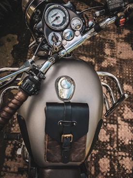 Close-up view of a motorcycle's fuel tank and dashboard, featuring a classic speedometer with a metallic finish. The handlebars and controls are visible, along with a leather strapped pouch attached to the fuel tank. The background includes a textured outdoor surface.