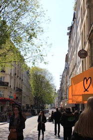 A bustling city street with tall historic buildings on either side, lined with trees that have lush green leaves. People walk along the street under the bright daylight, some carrying bags. An orange sign with a heart symbol stands out on the right side.