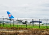 An airplane with the China Southern logo is taxiing on a runway, surrounded by a security fence with barbed wire at the top. The aircraft has a blue tail and is set against a grassy field and overcast sky.