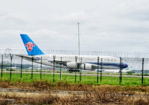 An airplane with the China Southern logo is taxiing on a runway, surrounded by a security fence with barbed wire at the top. The aircraft has a blue tail and is set against a grassy field and overcast sky.