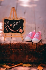A pair of pink baby shoes is placed next to a decorative mini chalkboard on a wooden surface. The chalkboard has ornate edges and bears the phrase 'Es Niña' written in elegant script with small floral and heart designs. The scene has a warm, soft lighting with a rustic outdoor ambiance indicated by fallen leaves.