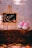 A pair of pink baby shoes is placed next to a decorative mini chalkboard on a wooden surface. The chalkboard has ornate edges and bears the phrase 'Es Niña' written in elegant script with small floral and heart designs. The scene has a warm, soft lighting with a rustic outdoor ambiance indicated by fallen leaves.