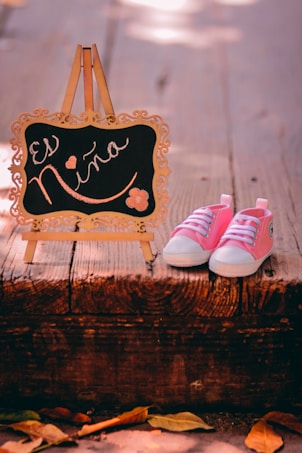A pair of pink baby shoes is placed next to a decorative mini chalkboard on a wooden surface. The chalkboard has ornate edges and bears the phrase 'Es Ni&ntilde;a' written in elegant script with small floral and heart designs. The scene has a warm, soft lighting with a rustic outdoor ambiance indicated by fallen leaves.
