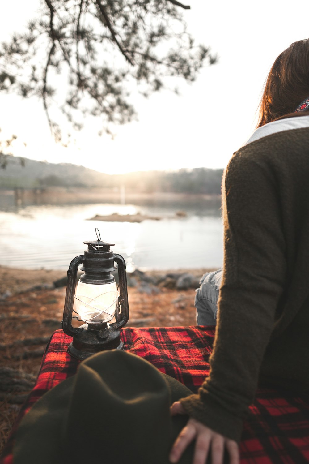 Mujer sentada en estera de picnic con linterna junto al lago