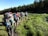 A group of hikers with large backpacks are walking in a single file along a narrow trail through a lush green meadow. The trail is surrounded by tall trees and the sky is clear. The hikers are dressed in outdoor attire, including hats and hiking boots.