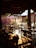 A cozy, open-air restaurant setting with wooden tables and chairs. Soft sunlight filters through the space, casting warm shadows and illuminating vibrant pink flowers in the background. The scene is peaceful and inviting, with decorative ceiling fans and hanging lights adding charm.