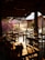 A cozy, open-air restaurant setting with wooden tables and chairs. Soft sunlight filters through the space, casting warm shadows and illuminating vibrant pink flowers in the background. The scene is peaceful and inviting, with decorative ceiling fans and hanging lights adding charm.