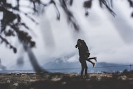 A couple embraces passionately in a misty, mountainous landscape. The silhouette of trees frames the scene, creating a sense of intimacy and seclusion. Overcast skies and the distant view of mountains enhance the dramatic atmosphere.