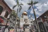 A grand mosque with a prominent golden dome is situated in a bustling urban area. Palm trees frame the foreground, while shops and market stalls line the streets. People are walking around, engaging in daily activities under a partly cloudy sky.