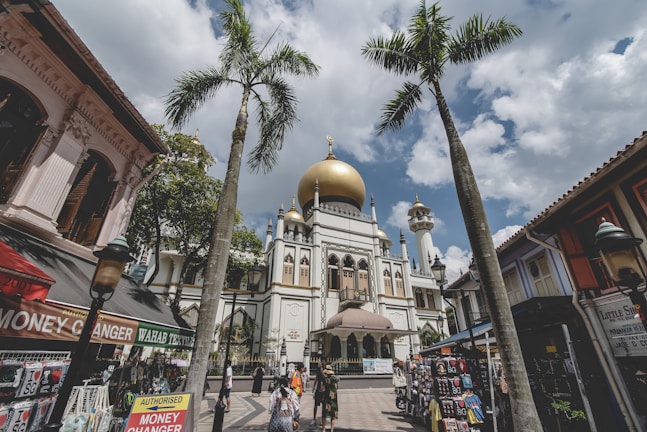 A grand mosque with a prominent golden dome is situated in a bustling urban area. Palm trees frame the foreground, while shops and market stalls line the streets. People are walking around, engaging in daily activities under a partly cloudy sky.