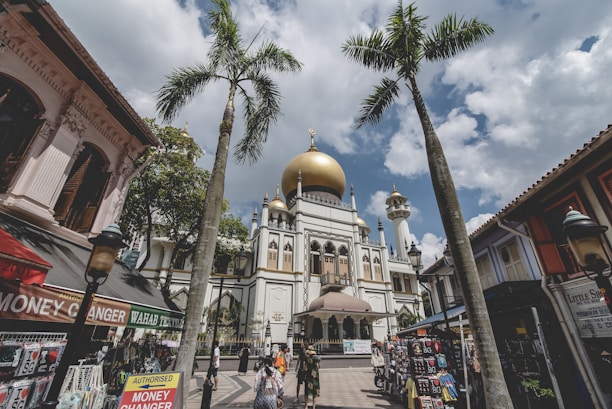 A grand mosque with a prominent golden dome is situated in a bustling urban area. Palm trees frame the foreground, while shops and market stalls line the streets. People are walking around, engaging in daily activities under a partly cloudy sky.