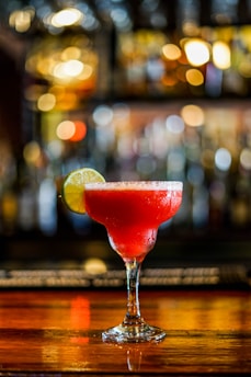 A vibrant red cocktail with a sugar-rimmed glass, garnished with a slice of lime, sits on a polished wooden bar. The background features a softly blurred array of warm, golden lights, creating a cozy and inviting atmosphere.