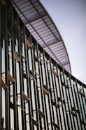 A modern building facade with tall vertical glass windows and metal frames. The structure has an overhang with a perforated metal design, creating a sleek and contemporary architectural look.