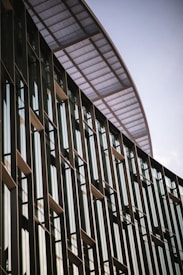 A modern building facade with tall vertical glass windows and metal frames. The structure has an overhang with a perforated metal design, creating a sleek and contemporary architectural look.