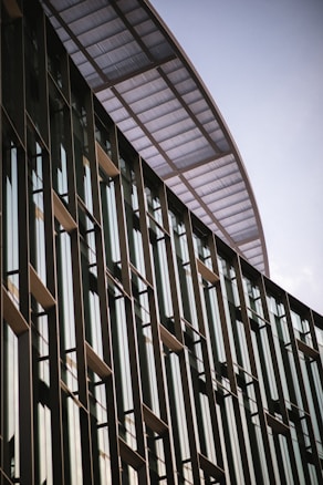 A modern building facade with tall vertical glass windows and metal frames. The structure has an overhang with a perforated metal design, creating a sleek and contemporary architectural look.