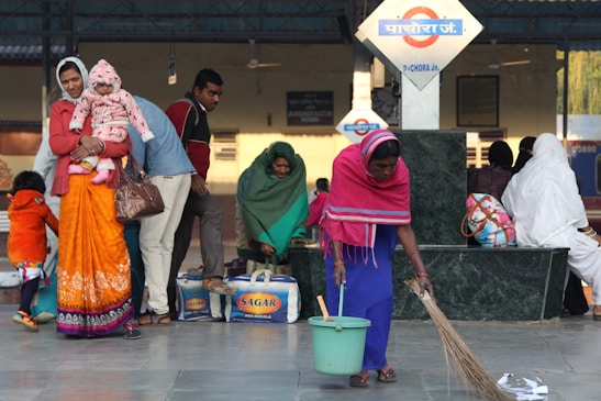A busy scene at a train station with multiple people visible. A woman in vibrant orange attire holds a child dressed in pink, while another woman in a pink shawl sweeps the floor. Nearby, several individuals sit and stand, some with luggage and bags. A sign in the background suggests this is Pachora Junction.