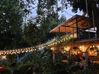 A cozy outdoor dining area set in a lush garden with string lights creating a warm and inviting atmosphere. The area features a pergola with a fabric awning, rustic brick arches, and various plants adding to the ambiance. People are seated under the pergola enjoying a meal.