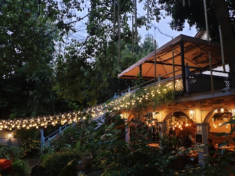 A cozy outdoor dining area set in a lush garden with string lights creating a warm and inviting atmosphere. The area features a pergola with a fabric awning, rustic brick arches, and various plants adding to the ambiance. People are seated under the pergola enjoying a meal.