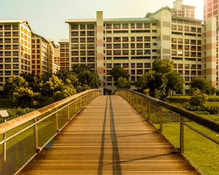 A pedestrian bridge with metal railings leads towards a cluster of modern residential buildings. The sunlight casts elongated shadows across the wooden planks of the bridge. Trees and greenery surround the area, adding a natural contrast to the urban setting.
