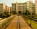 A pedestrian bridge with metal railings leads towards a cluster of modern residential buildings. The sunlight casts elongated shadows across the wooden planks of the bridge. Trees and greenery surround the area, adding a natural contrast to the urban setting.