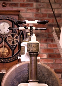 An antique mechanical device with a metal handle and labeled 'Lehmann Dresden' is prominently displayed. Behind it, there is a clock and gears mounted on a brick wall, suggesting an industrial or workshop setting.